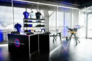 Two black bars with apparel displayed: purple, white, and black t-shirts as well as branded black belt bags. To the right, a t-shirt printing press.