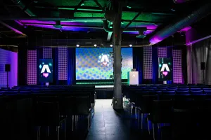 View from the back of the dark empty keynote presentation area. Rows empty black chairs. A vibrant large LED screen with the Astro Together logo on it. Two smaller portrait-oriented screens adorn the sides of the stage, also with the Astro Together logo.