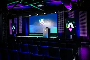 View from the back of the dark empty keynote presentation area. Rows empty black chairs. A vibrant large LED screen with the Astro Together logo on it. Two smaller portrait-oriented screens adorn the sides of the stage, also with the Astro Together logo.