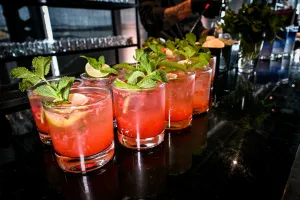 Half a dozen pink cocktails garnished with mint and lemon atop a black bar.