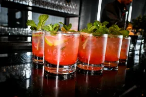 Half a dozen pink cocktails garnished with mint and lemon atop a black bar.