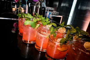 Half a dozen pink cocktails garnished with mint and lemon atop a black bar.