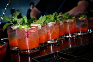 Half a dozen pink cocktails garnished with mint and lemon atop a black bar.