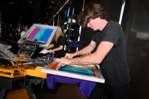 A man using the t-shirt printing press to print a purple t-shirt for a guest.