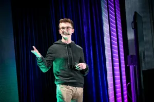 A close up of Ben Holmes on stage gesturing to and looking at the LED screen.