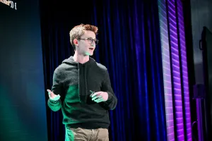 A close up of Ben Holmes on stage gesturing to the LED screen while looking at the audience.