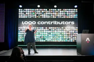 Sarah Rainsberger speaking from the left side of the stage, talking about how Docs has had 1,000 contributors to-date.