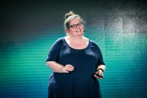 Sarah Rainsberger speaking on stage in front of a green and black LED screen.