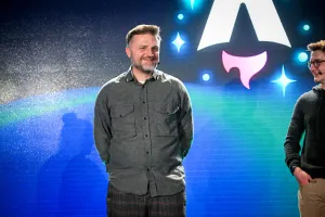 Closeup of Matthew Phillips standing with other keynote speakers on stage. His hands are behind his back and he is smiling at the audience. Off to the right, Ben Holmes is smiling at him.