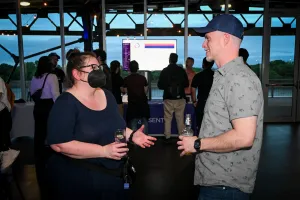 Sarah Rainsberger, wearing a black mask, chats with a guest. The Sentry booth and TV screen can be seen behind them.